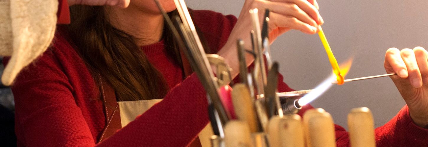 Mains d'une stagiaire utilisant un outil de modelage métallique pour aplatir une perle de verre sous le chalumeau.