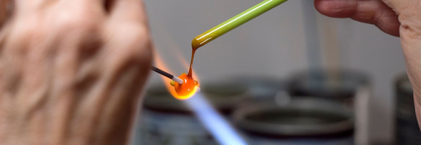 Deux personnes côte à côte manipulant des mandrins et des baguettes de verre colorées devant la flamme.