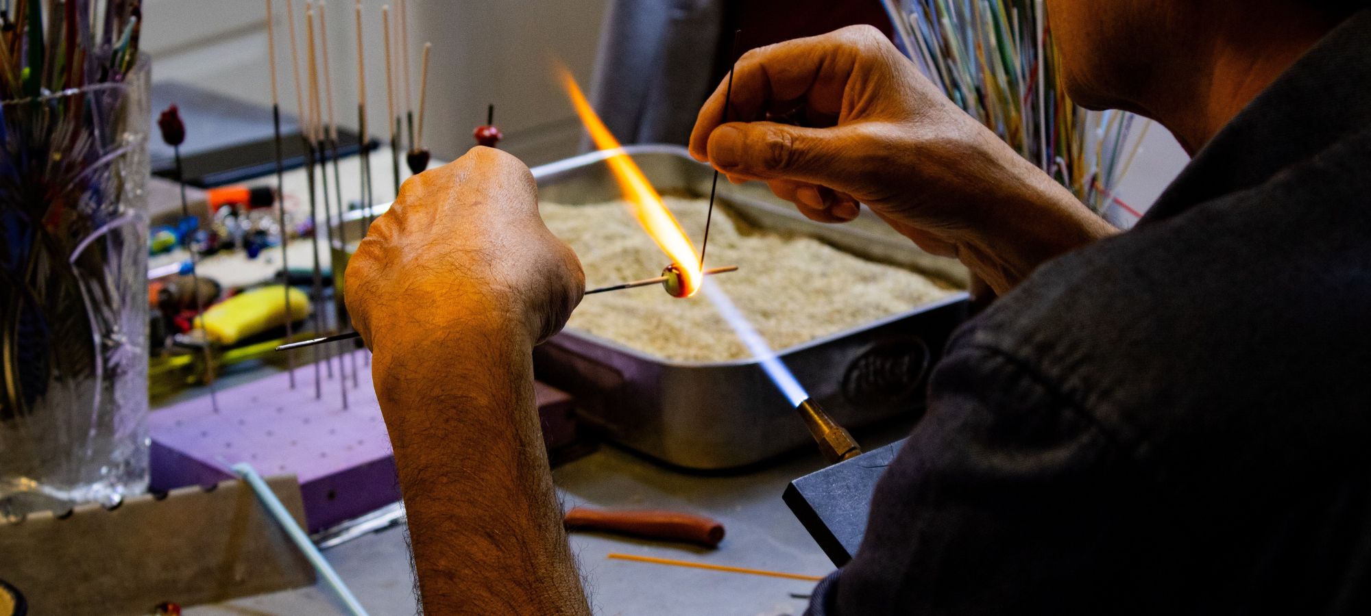 Atelier créatif verre en fusion Stagiare manipulant un fil de verre et un mandrin devant un chalumeau professionnel de perlière.